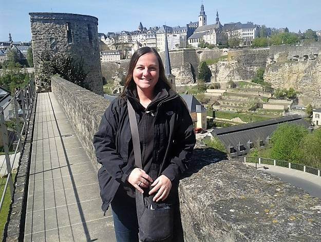 a girl dressed in a black coat, black fleece and blue jeans on an old castle wall with a valley with trees and grass and buildings on the land above