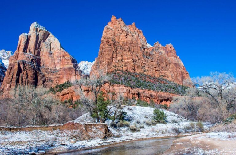 Snow-dusted landscape of Zion National Park featuring towering red rock cliffs beneath a vivid blue sky, with a shallow river winding through a foreground of bare winter trees and light snow cover.