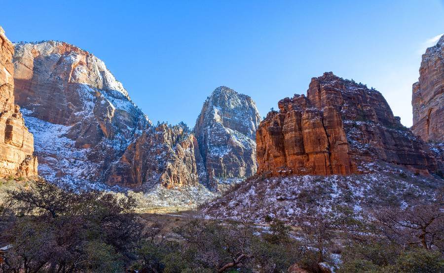 Scenic winter landscape of Zion National Park featuring towering red rock cliffs partially covered in snow, with clear blue skies above and a dusting of snow on the trees and valley floor below.