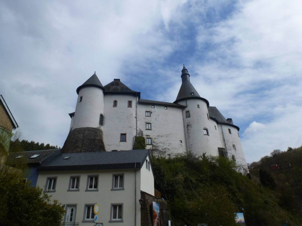 View of Clervaux Castle towering above the hillside in Clervaux, Luxembourg, with its white walls, round defensive towers, and dark slate cone roofs rising behind nearby village houses under a cloudy blue sky.