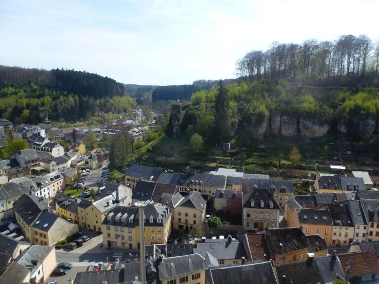 Elevated view of the village of Larochette in Luxembourg, showing tightly clustered houses with slate roofs, narrow streets, and surrounding green hills and forested cliffs under a bright sky.