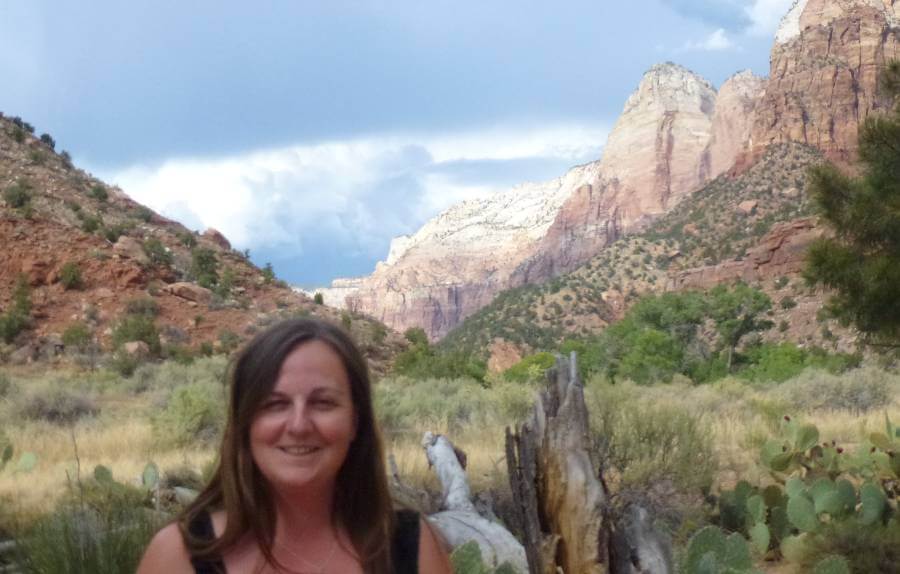 A woman smiling in the foreground of a desert landscape in Zion National Park, with red rock cliffs, spiky cactus plants, dry grasses, and dramatic clouds in the background. The towering sandstone formations are lit by sunlight, contrasting with the darker storm clouds behind.