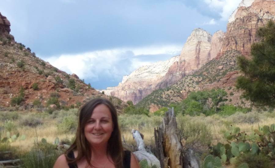 Woman smiling in the foreground of a desert valley in Zion National Park, with red and white sandstone cliffs rising behind her under dramatic gray clouds. Desert grasses, prickly pear cactus, and scattered shrubs fill the landscape beneath the towering canyon walls.