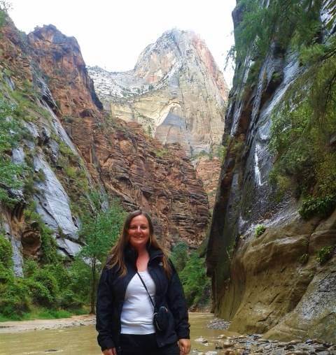 Woman standing in a narrow canyon with towering red and cream sandstone cliffs rising steeply on both sides. She is smiling at the camera, wearing a black jacket and light-colored top, with lush greenery and a shallow rocky stream running through the canyon beneath a bright sky above.